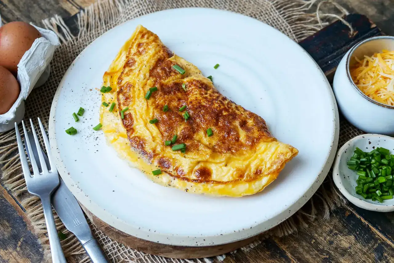 Goldbraunes Omelett mit K&auml;se und Schnittlauch, perfekt f&uuml;r ein schnelles Omelett Rezept. Daneben Eier, K&auml;se und Schnittlauch.