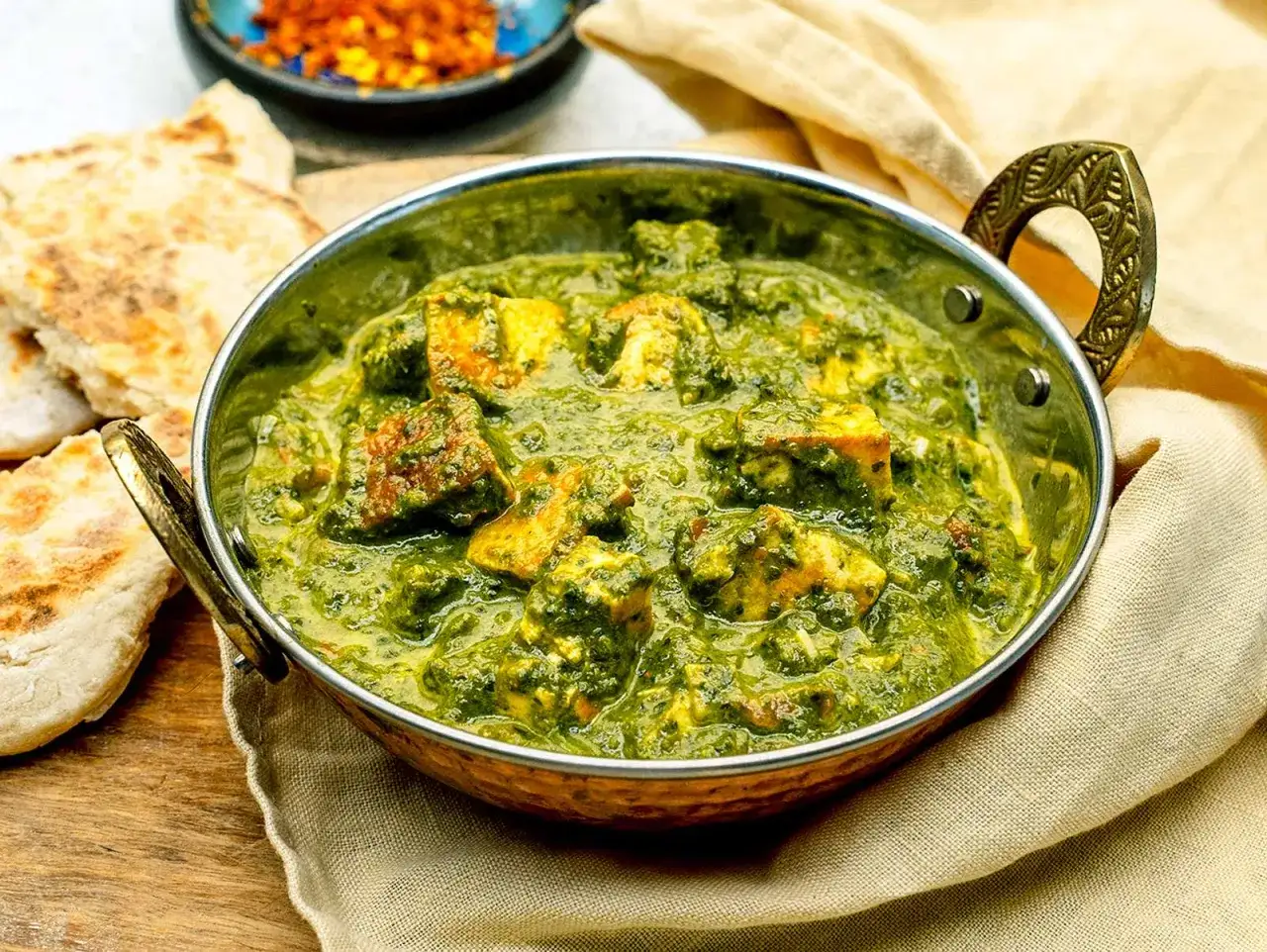 Ein k&ouml;stliches Palak Paneer in einer Metallschale, serviert mit Fladenbrot und Chiliflocken.