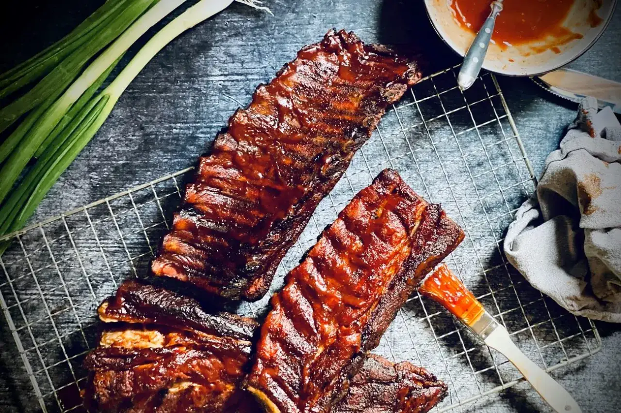 Saftige spare ribs, glasiert mit BBQ-Sauce, liegen auf einem Grillrost. Fr&uuml;hlingszwiebeln und eine Sch&uuml;ssel Sauce daneben deuten auf ein k&ouml;stliches Mahl hin.