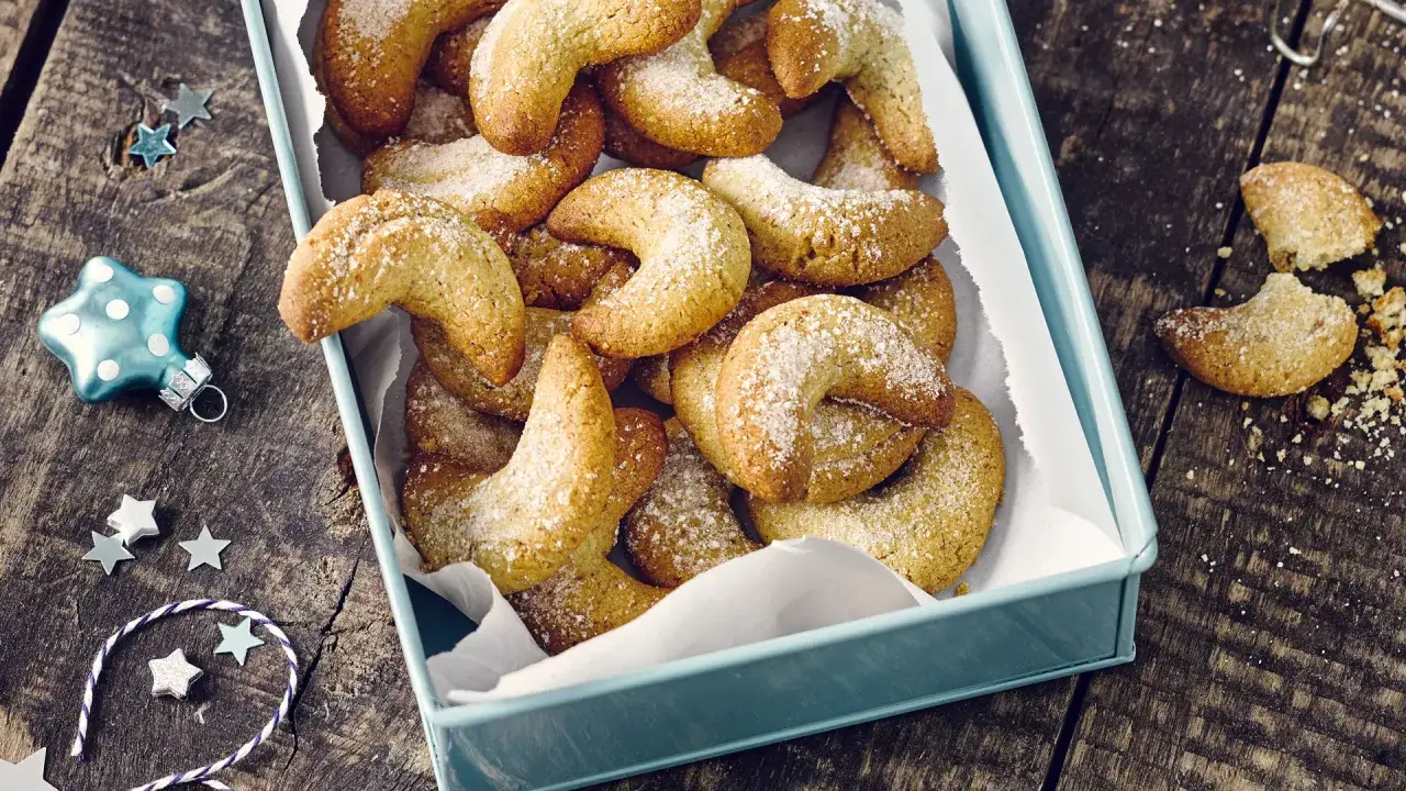 Ein Blech voller goldbrauner Vanillekipferl, best&auml;ubt mit Puderzucker, liegt auf einem rustikalen Holztisch. Weihnachtliche Deko wie Sterne und eine blaue Christbaumkugel schm&uuml;cken die Szene.