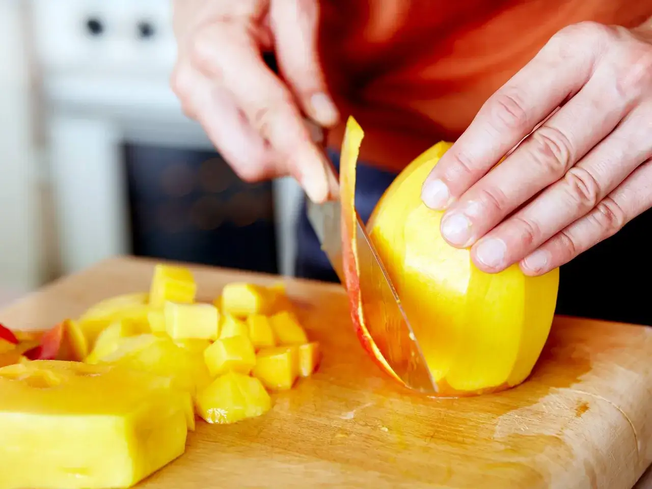 H&auml;nde sch&auml;len eine Mango auf einem Holzbrett. Daneben liegen bereits gew&uuml;rfelte Mangost&uuml;cke. Man kann Mango mit Schale essen, aber hier wird sie gesch&auml;lt.