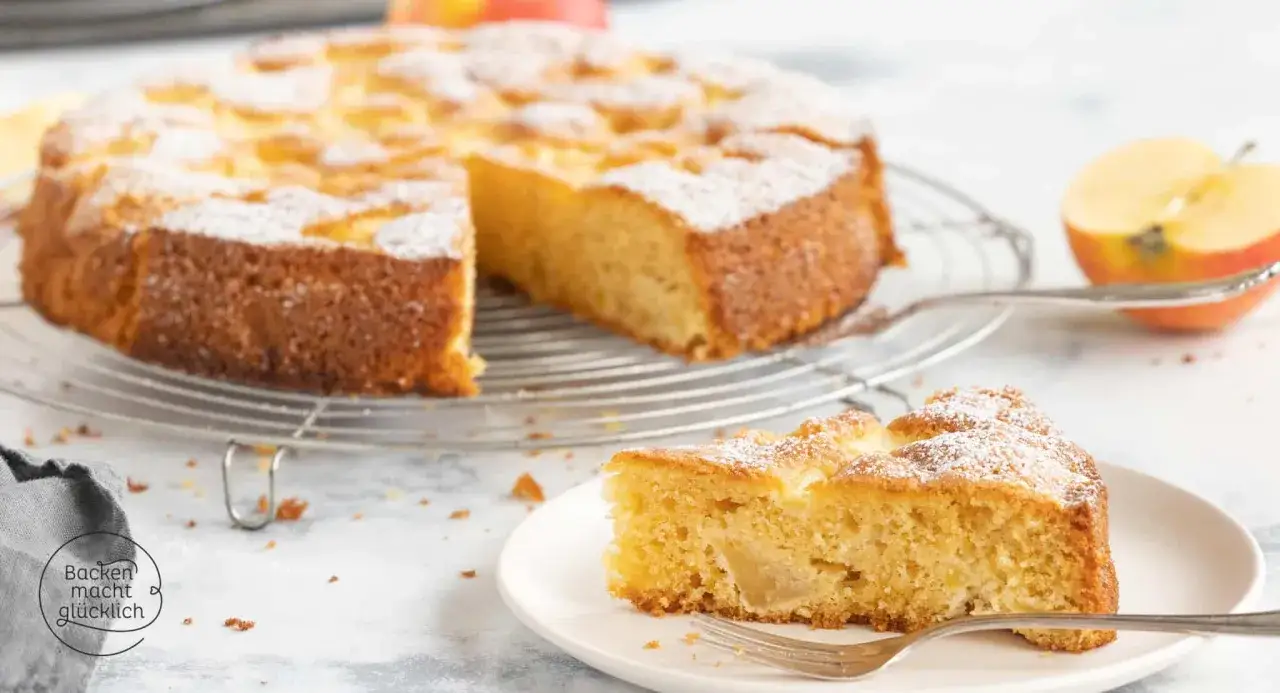 Ein saftiger, glutenfreier Apfelkuchen, best&auml;ubt mit Puderzucker, wartet auf einem Kuchengitter. Ein St&uuml;ck auf einem Teller mit Gabel daneben.