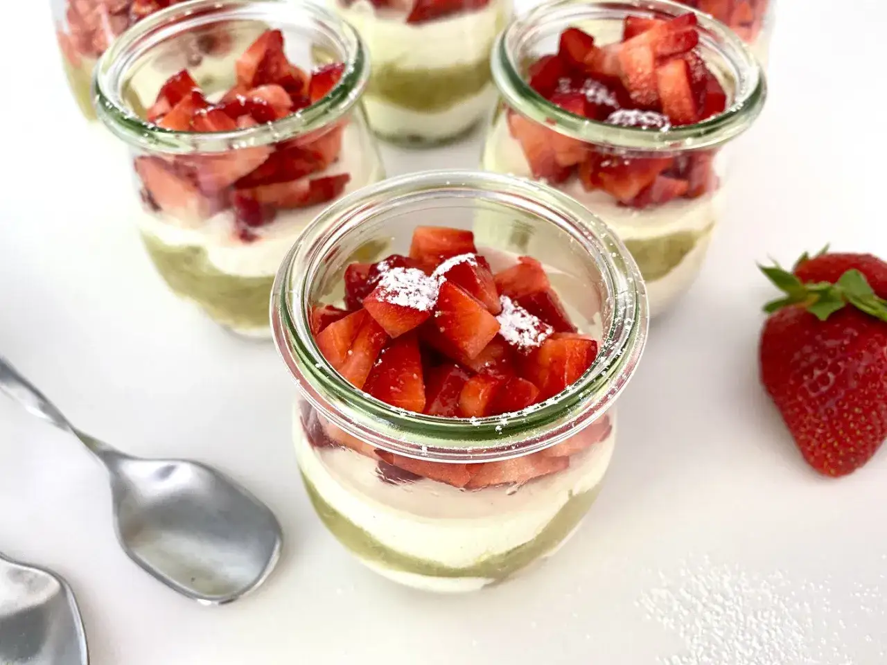 Mehrere Gl&auml;ser mit Schichten aus gr&uuml;nem Pudding, wei&szlig;er Creme und frischen Erdbeeren. Ein Hauch von Rhabarberkompott k&ouml;nnte die s&uuml;&szlig;e Note abrunden.