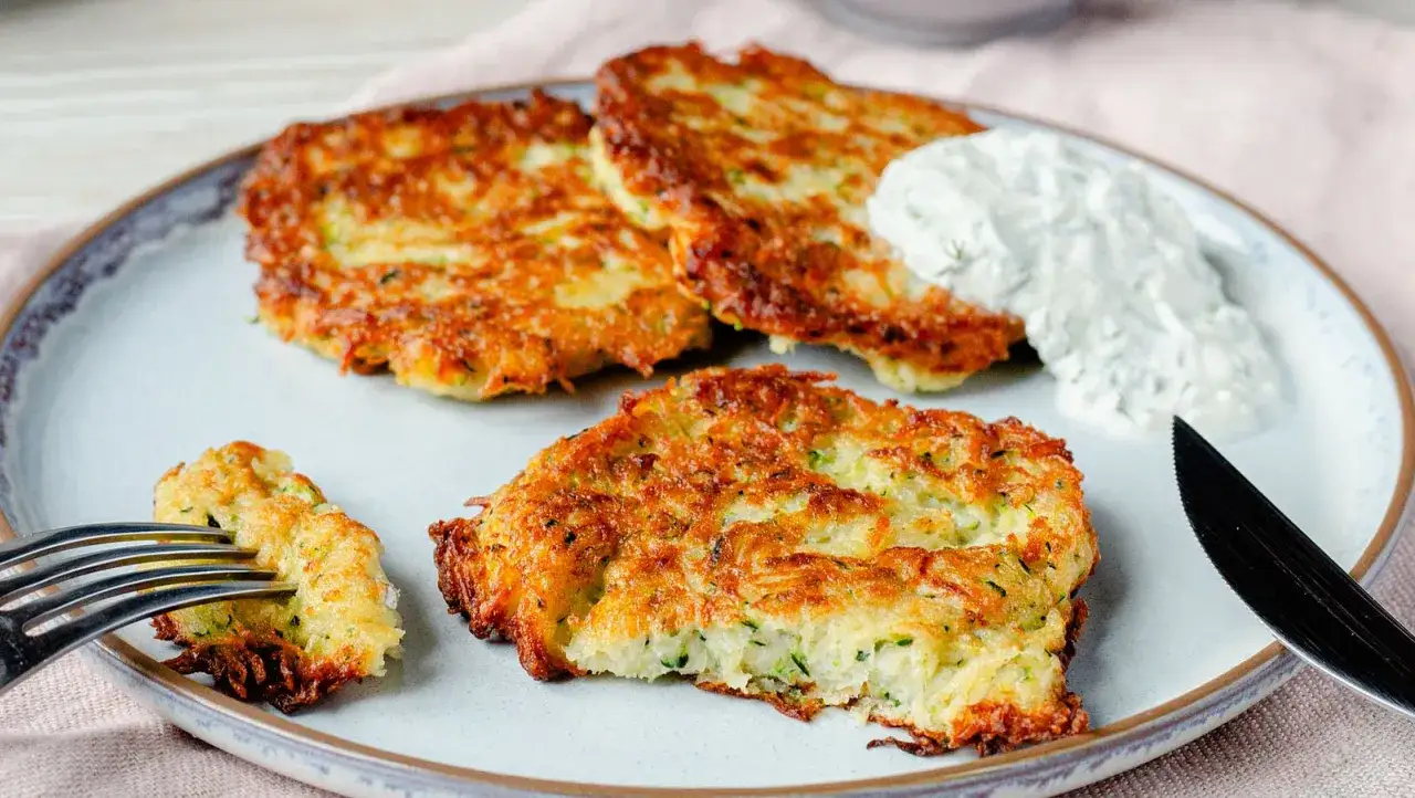 Goldbraune zucchini karotten puffer auf einem Teller mit Dip. Eine Gabel nimmt ein Stück.