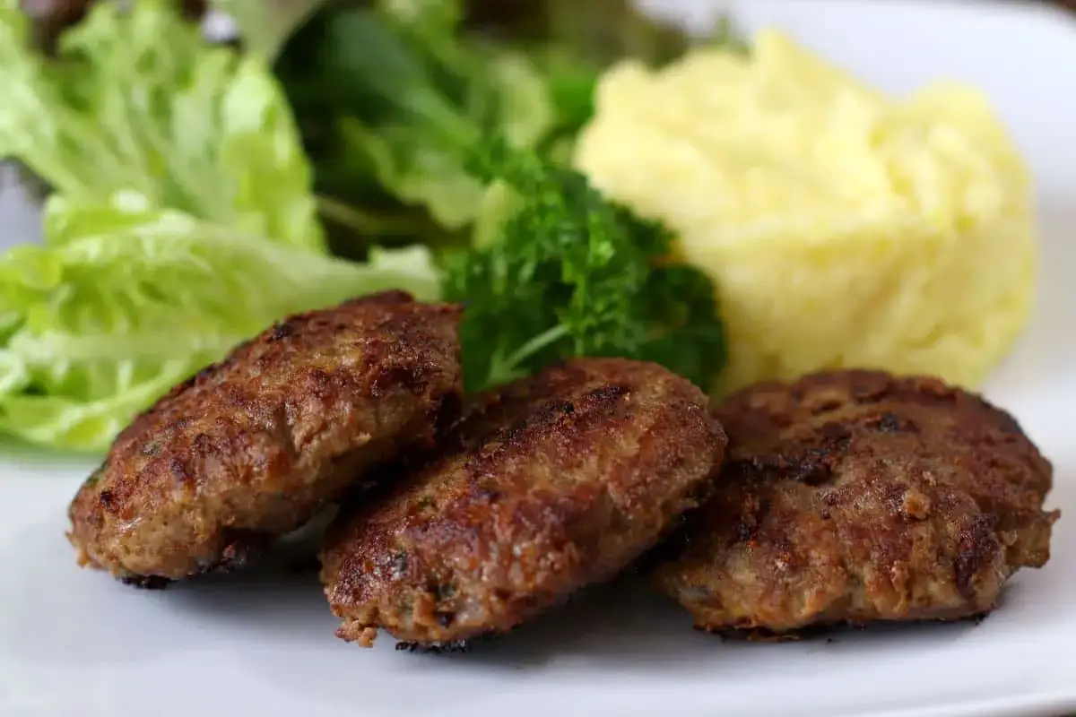Drei goldbraune Fleischpflanzerl liegen auf einem Teller neben Kartoffelbrei und Salat. Ein leckeres Fleischpflanzerl Rezept zum Nachkochen.