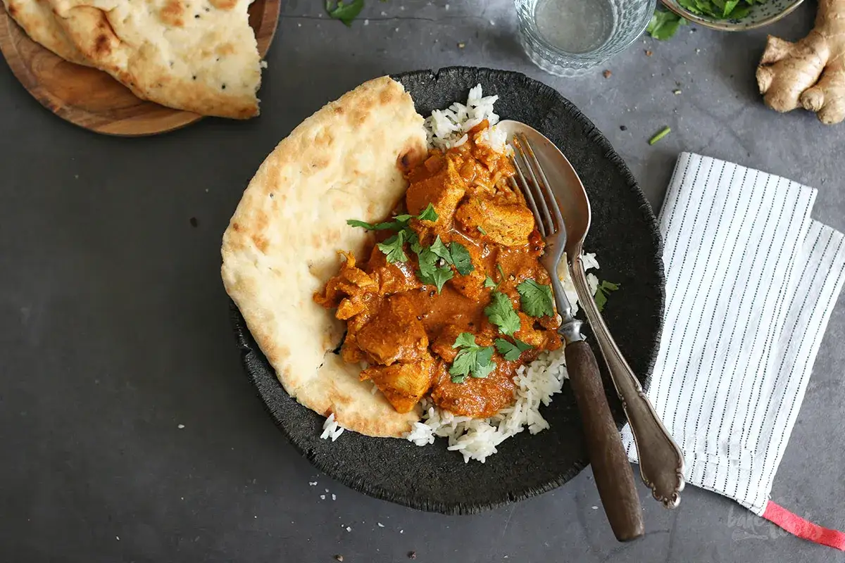 Ein Teller mit cremigem Butter Chicken, Reis und Naan-Brot, garniert mit Koriander.