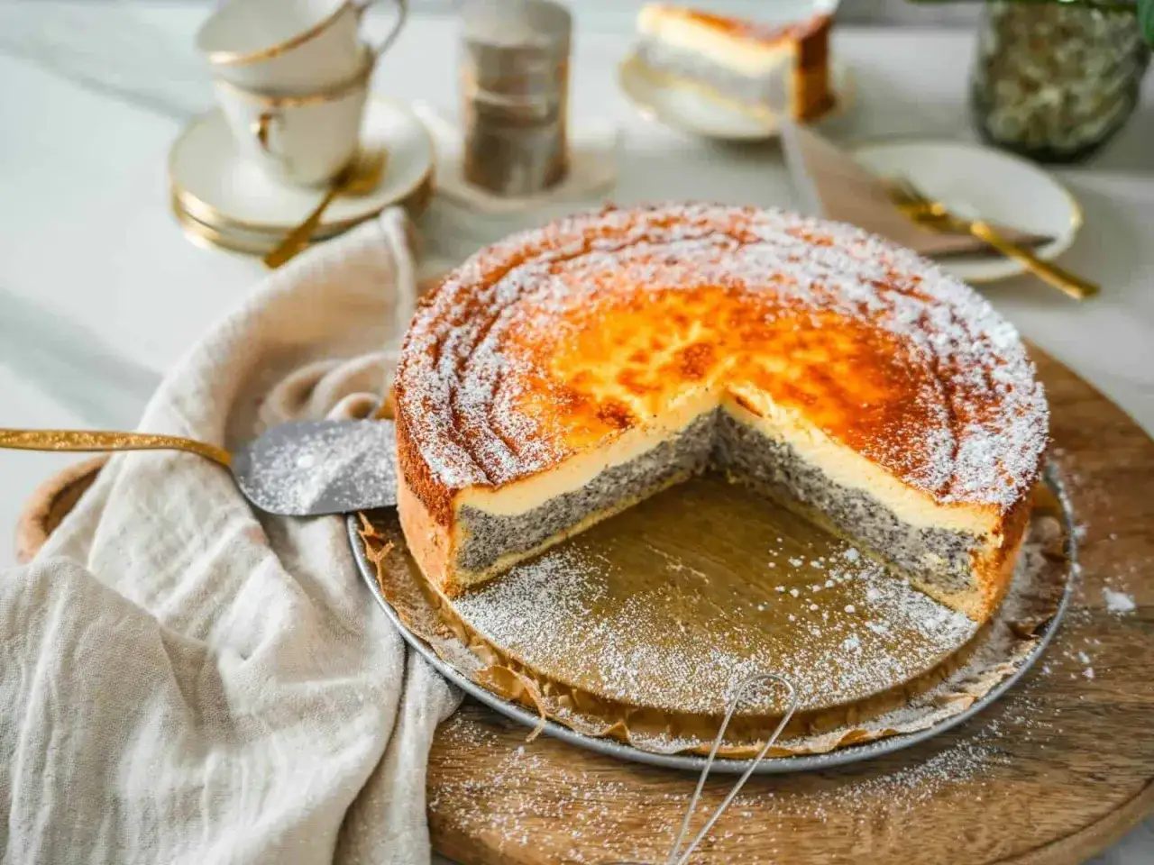 Ein köstlicher Mohn Schmand Kuchen, bestäubt mit Puderzucker, wartet darauf, angeschnitten zu werden. Daneben liegen Kaffeetassen.