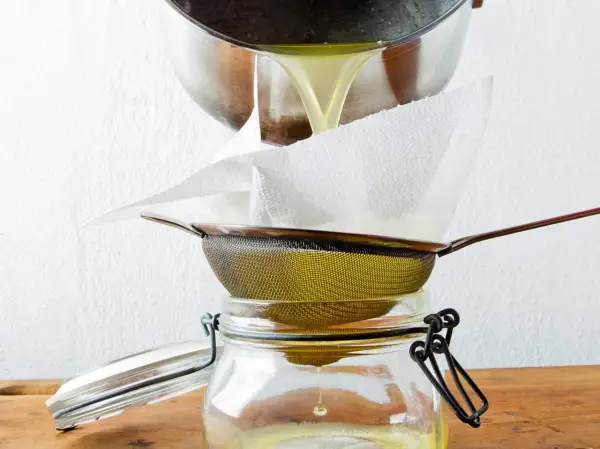 Flüssiges Ghee wird durch ein Sieb mit Küchenpapier in ein Glas gefiltert. So gelingt das Ghee selber machen.
