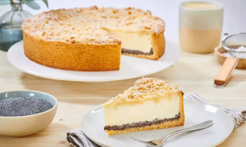 Ein köstlicher Mohn Schmand Kuchen mit Streuseln und Puderzucker. Daneben eine Schale Mohn und eine Tasse Kaffee.